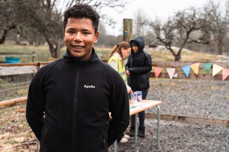 Ung person står utomhus och ler mot kameran, med volontärer och ett bord i bakgrunden.