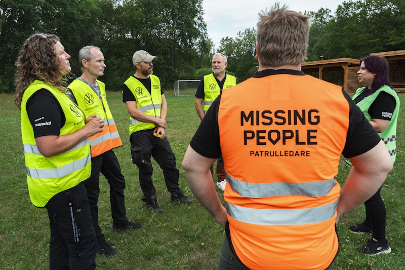 Volontärer från Missing People samlas utomhus för genomgång, flera bär reflexvästar.